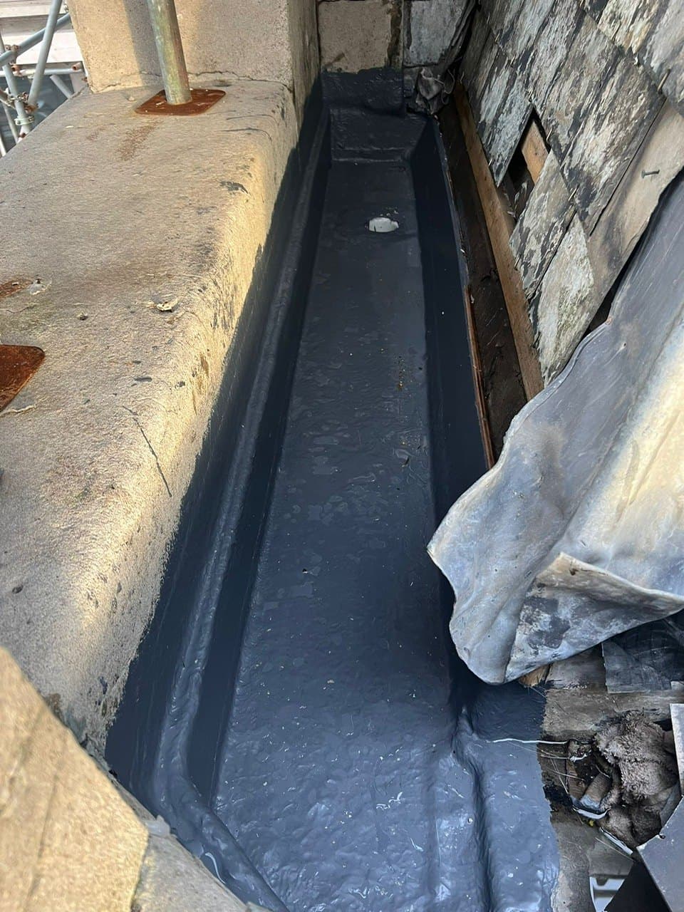 Black liquid-applied membrane in parapet box gutter beside chimney — gutter re-line work.